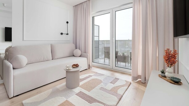 A modern living room with a sofa, a coffee table, a large window with curtains, and a view of the city. - Spacious Apartment | Balcony and Private Parking in Garage (Łódź)