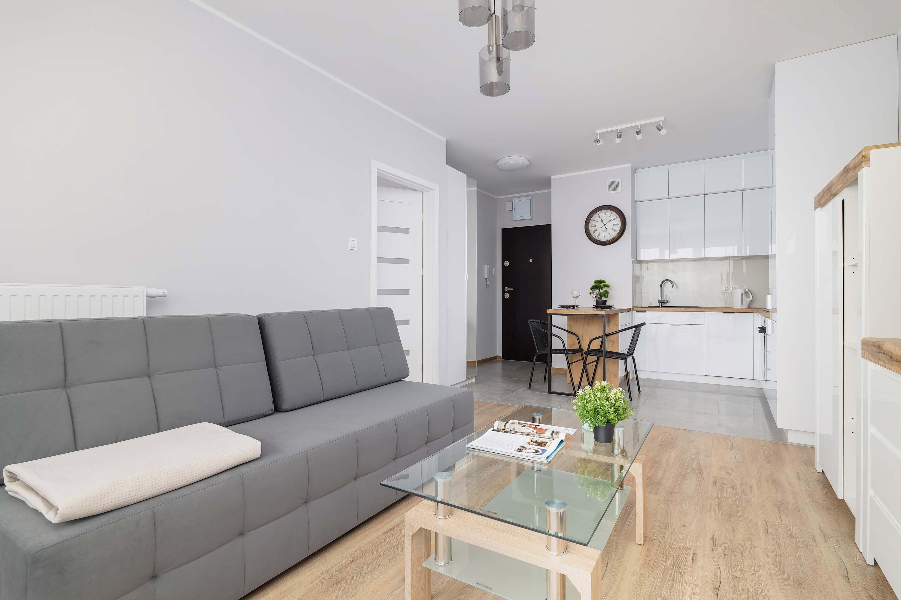 Un salon moderne avec un canapé gris, une table basse en verre et une cuisine en arrière-plan.