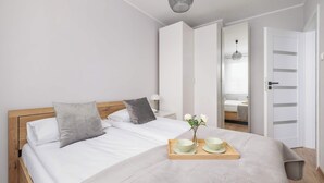 Un lit bien fait, avec du linge de lit blanc, une tête de lit en bois et un plateau sur lequel se trouvent deux bols et un vase de fleurs.