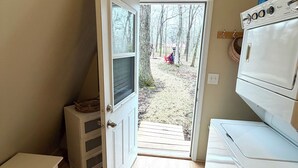 Une buanderie équipée d’un lave-linge et d’un sèche-linge, d’un petit meuble blanc et d’une porte donnant sur un espace extérieur avec des arbres et une terrasse en bois.