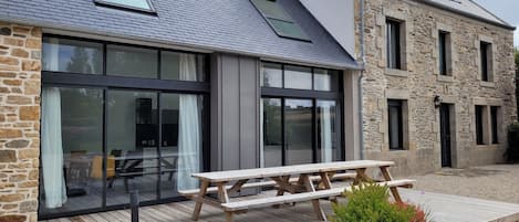 Ein modernes Haus mit Fassade aus Stein und Metall, großen Glasfenstern und einer Holzterrasse mit Picknicktisch.