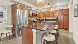 A modern kitchen with wooden cabinets, stainless steel appliances, a granite countertop, and bar stools. - Ember & Ash | Designer Home w/ King Beds, Games (Fort Collins)