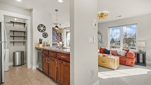 A modern kitchen with wooden cabinets, a stainless steel dishwasher, and a built-in oven. There is a dining area with a red sofa and a coffee table. The room has large windows providing natural light. - The Revive Loft | Balcony, Workspace & Bikes (Fort Collins)
