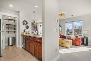 A modern kitchen with wooden cabinets, a stainless steel dishwasher, and a built-in oven. There is a dining area with a red sofa and a coffee table.