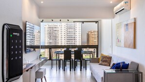 A modern living room with a sofa, dining table, and chairs, a built-in air conditioner, and a view of high-rise buildings. - Barra Max - Praia e Conforto. (Rio de Janeiro)