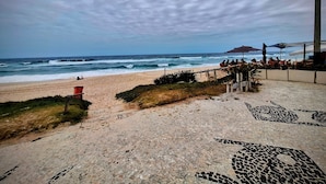 A beach with a paved walkway leading to the shore, a red trash bin, and umbrellas providing shade. - Barra Max - Praia e Conforto. (Rio de Janeiro)