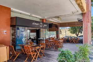 Un café en plein air avec des tables et des chaises en bois, une enseigne indiquant « cinco 2 zero », et une vue sur des voitures garées et un arbre.