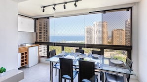 A modern dining area with a glass table, chairs, and a city view through large windows. - Barra Max - Praia e Conforto. (Rio de Janeiro)
