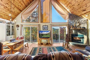 A spacious living room with a high ceiling, wooden beams, a stone fireplace, a television, and a large window with a mountain view. - Green Forest Lodge - Pet Friendly (Murphy)