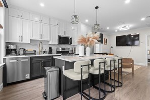 A modern kitchen with white cabinets, stainless steel appliances, and a central island with bar stools. - Modern 3BR Near NYC + MetLife • Sleeps 8 + Parking (Union City)