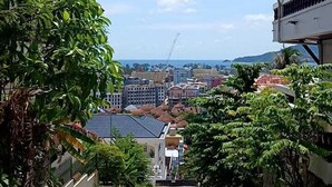 A paved pathway leading to a building with a sloped roof, surrounded by lush greenery and a clear view of a distant cityscape. - Aksarai Hotel Patong  (Patong)