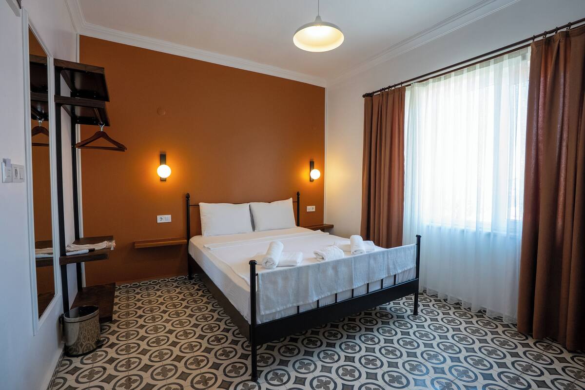 A hotel room with a bed, two bedside tables, a hanging coat rack, a patterned floor, and a window with curtains.