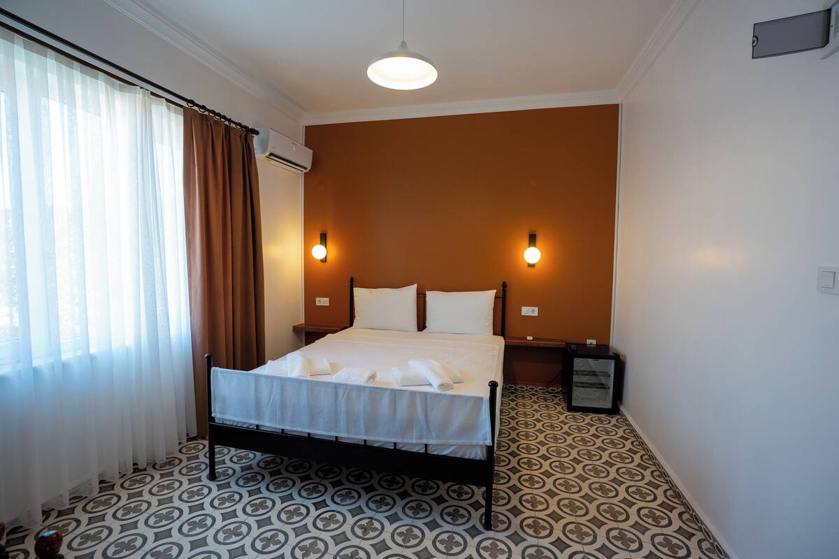 A hotel room with a bed, curtains, patterned floor, wall-mounted lights, and an air conditioning unit.