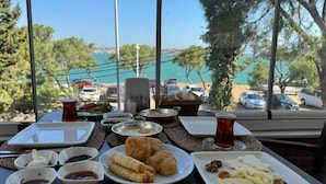 Una mesa dispuesta con alimentos para el desayuno, incluyendo cruasanes, queso y nueces, con vista al mar y coches estacionados al fondo.