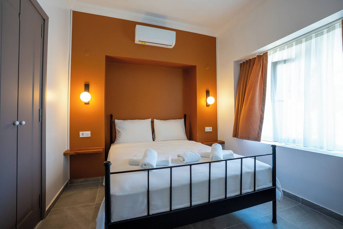 A hotel room with a bed, two wall-mounted lights, a window with curtains, and an air conditioning unit.