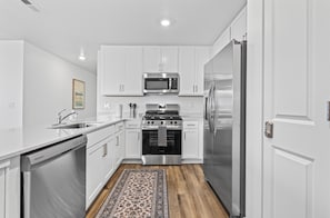 A modern kitchen with white cabinetry, stainless steel appliances, and a patterned rug. - NEW 4BR by Ford Center • Pool Access + Fenced Yard (Nampa)
