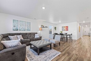 A modern living room with a sectional sofa, ottoman, and patterned rug. There is a dining area with a table and chairs, and a kitchen in the background. - NEW 4BR by Ford Center • Pool Access + Fenced Yard (Nampa)