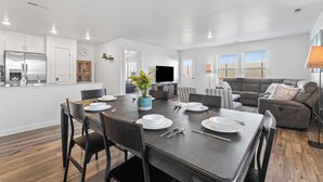 Une cuisine moderne avec des meubles blancs, un coin repas doté d’une table en bois foncé et un canapé gris dans le salon.