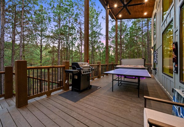 A wooden deck with a grill, table tennis table, and a view of a forest. - Ping-Pong Table | Corn Hole | EV Charger | Hot Tub (Broken Bow)