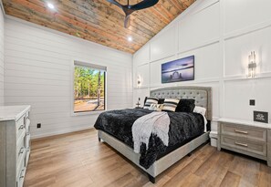 A bedroom with a wooden ceiling, a large bed with a grey upholstered headboard, a window with a view of trees, and a wall-mounted picture. - Ping-Pong Table | Corn Hole | EV Charger | Hot Tub (Broken Bow)