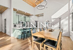 A modern dining area with a wooden table and chairs, a kitchen in the background, and a chandelier. - Ping-Pong Table | Corn Hole | EV Charger | Hot Tub (Broken Bow)