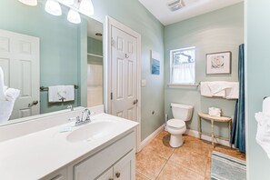 Casa de banho com uma sanita, um lavatório branco e um espelho. Tem uma janela com cortina e um porta-toalhas com toalhas.