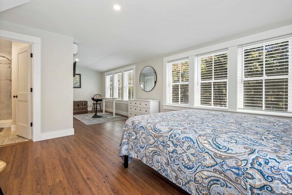 A bedroom with a large bed, a desk, and a chair. There are windows with blinds and a door leading to another room.