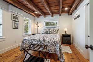 Une chambre avec un plafond en bois, un lit recouvert d’une literie à motifs floraux, une table de chevet, une lampe et une fenêtre avec des stores.