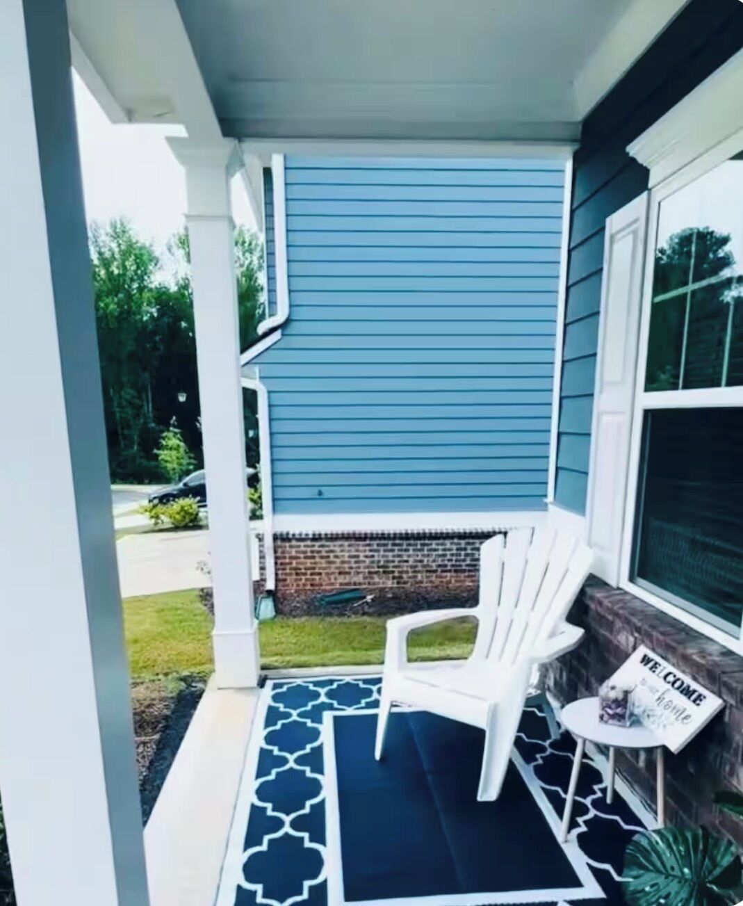 A porch with a blue siding house, a white chair, a small table with a 'Welcome' sign, and a patterned rug.