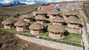 A cluster of thatched-roof huts with stone walls, arranged in a semi-circular pattern, surrounded by a stone fence, with a mountainous backdrop. - Pachamama Hampina Wasi Centro Ancestral - All Inclusive (Maras)