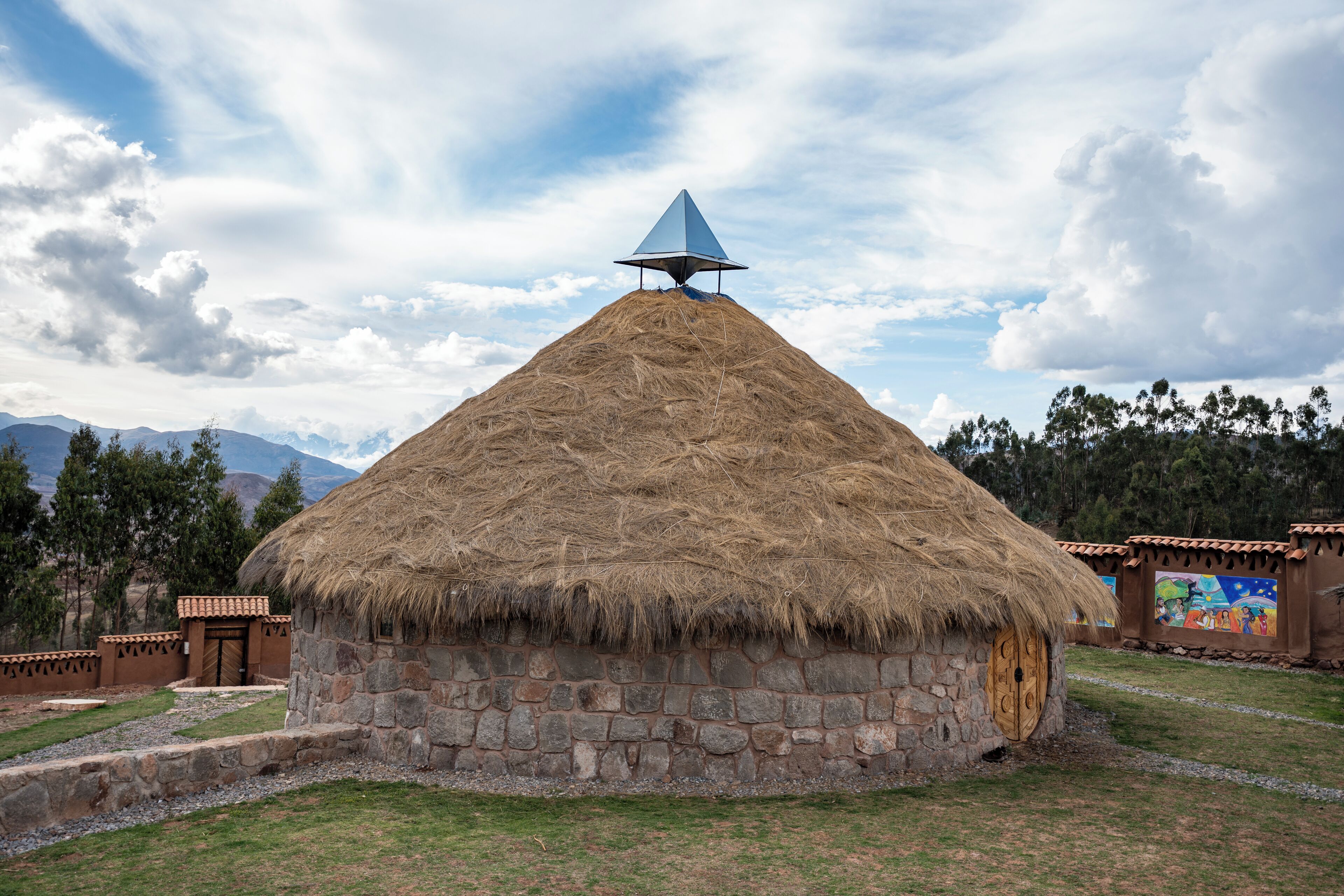 Eine traditionelle, reetgedeckte Hütte mit Steinunterbau, umgeben von einer angelegten Grünfläche mit Wegen und einem Wandgemälde.