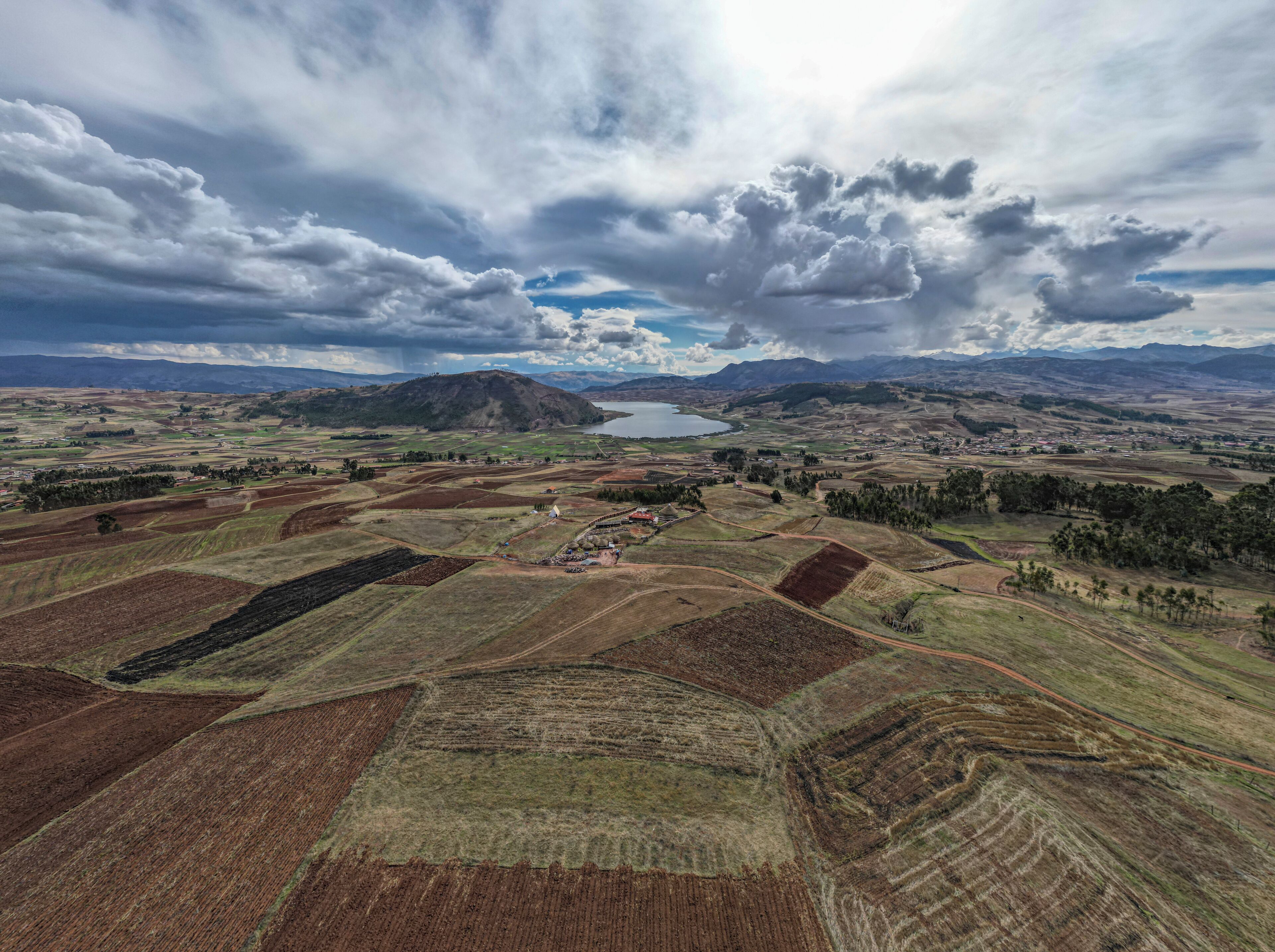 A panoramic view of a rural landscape with terraced fields, a small body of water, and distant mountains under a cloudy sky.