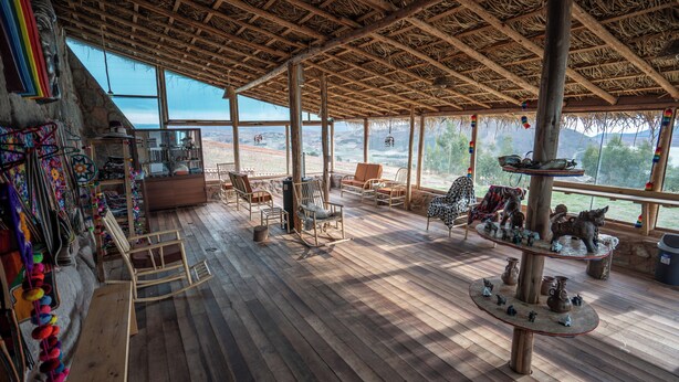 A spacious wooden deck with multiple seating areas, a central table with decorative items, and large windows offering views of the surrounding landscape. - Pachamama Hampina Wasi Centro Ancestral - All Inclusive (Maras)