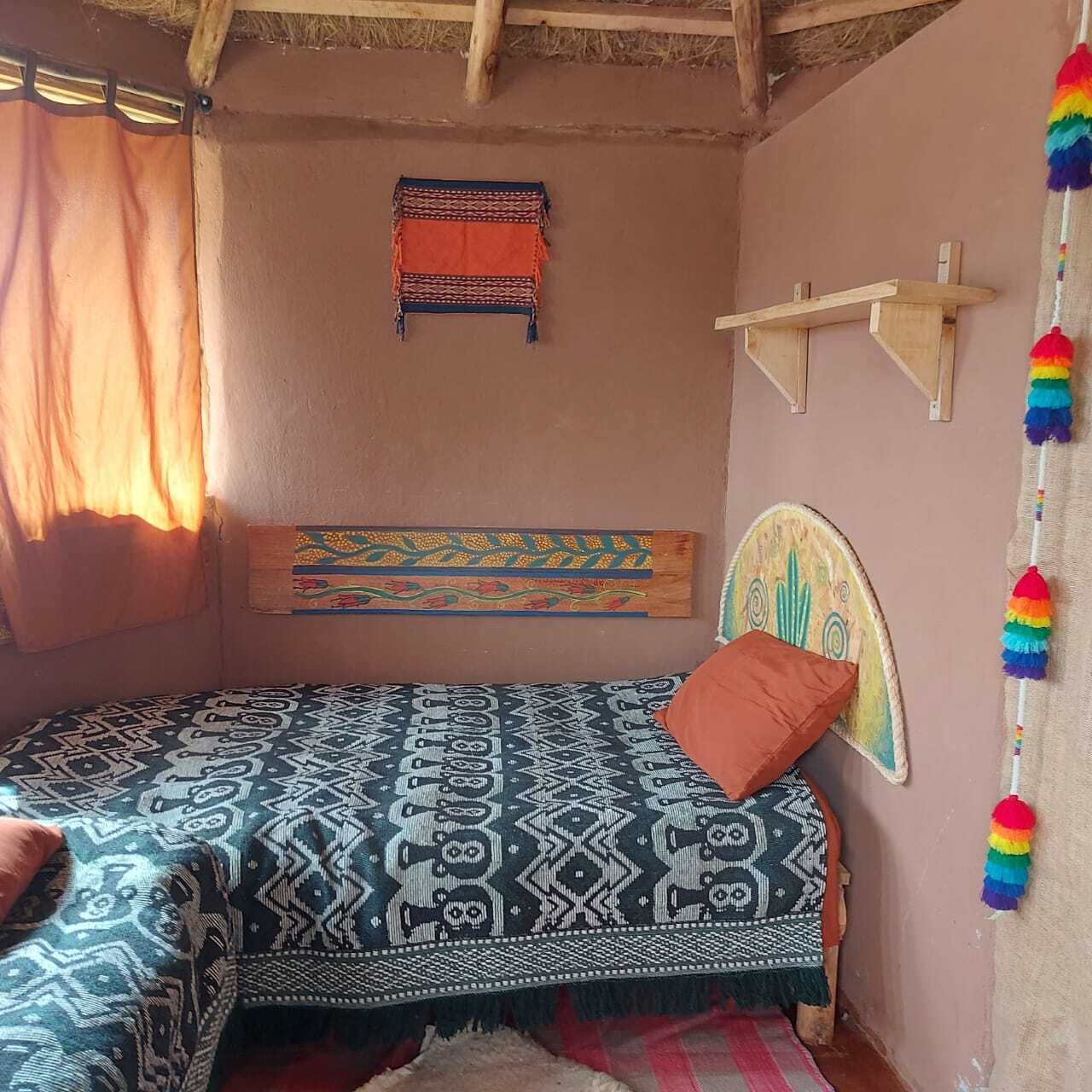 A small, cozy room with a bed, a window with curtains, a wall-mounted shelf, and a colorful hanging decoration.