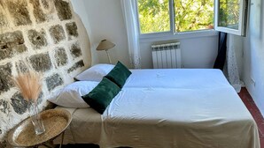 A bedroom with a stone wall, a bed with white and green bedding, a small table with a vase, and a window with a view of trees.