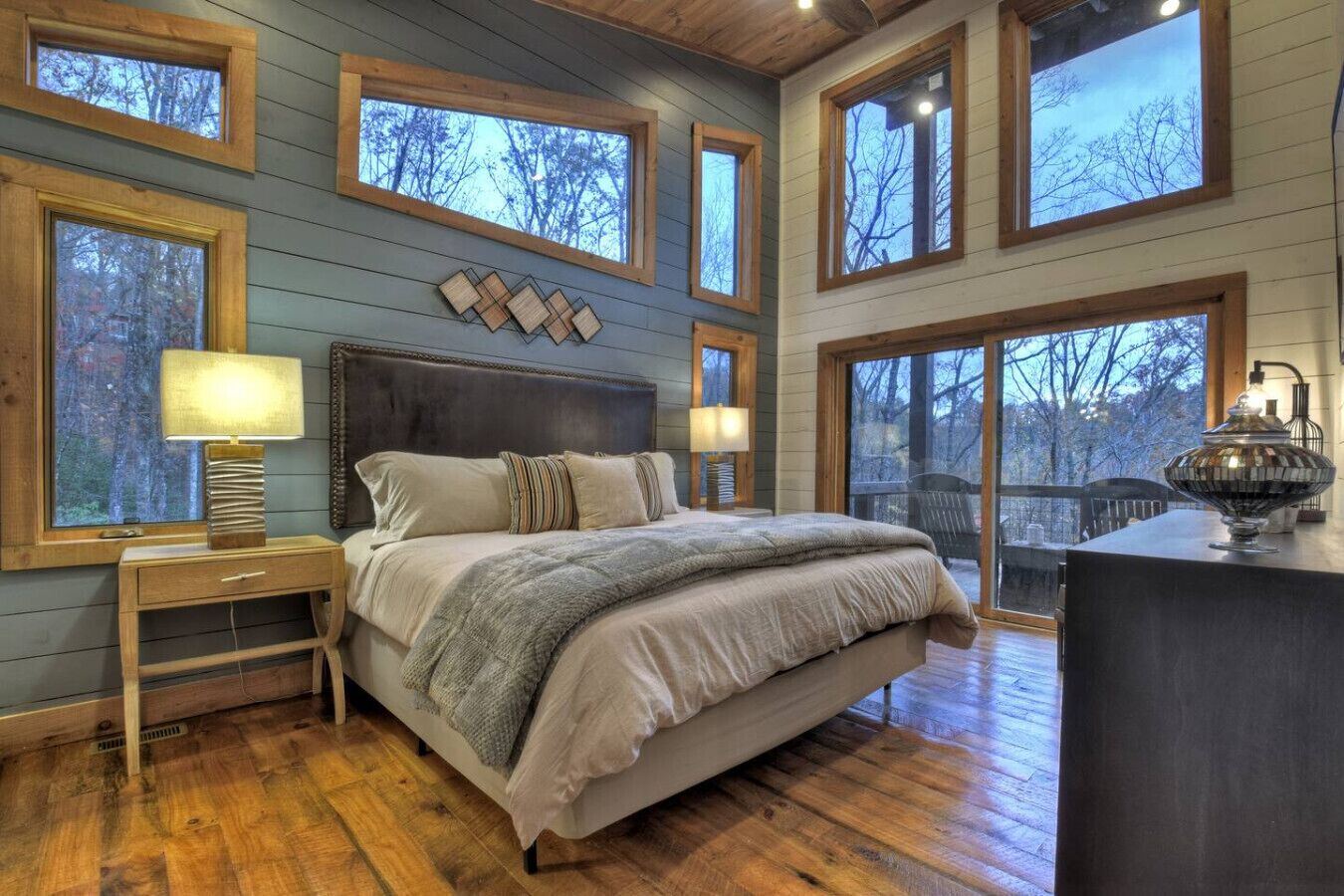 Un dormitorio con una cama grande, cabecera de madera, varias ventanas y vistas a árboles.