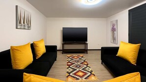 Una sala de estar con dos sofás negros, cojines amarillos, un televisor de pantalla plana sobre un mueble bajo, una alfombra estampada y obras de arte en la pared.