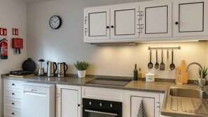 Una cocina con armarios blancos, un reloj, una cafetera, un hervidor, un tostador, una planta y diversos utensilios de cocina.