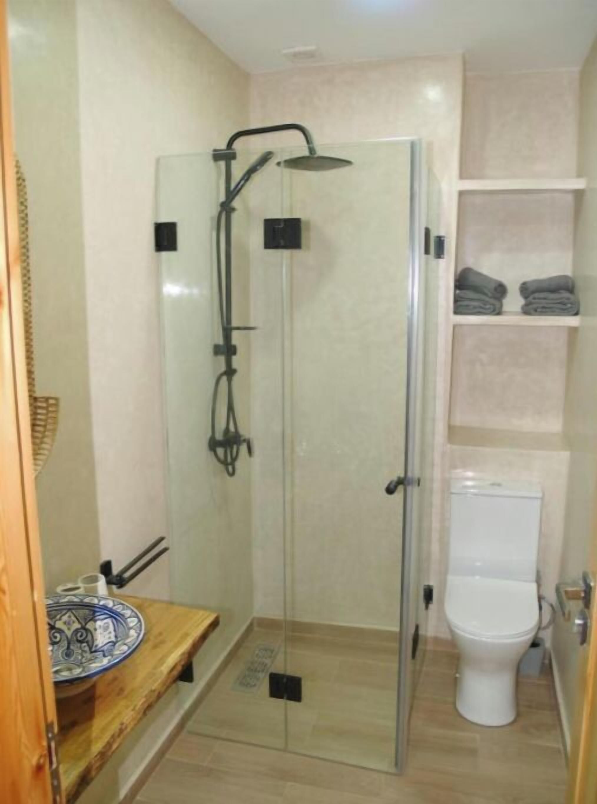 Un box de ducha con paredes de cristal, un cabezal de ducha de mano, un inodoro y un estante con toallas dobladas.