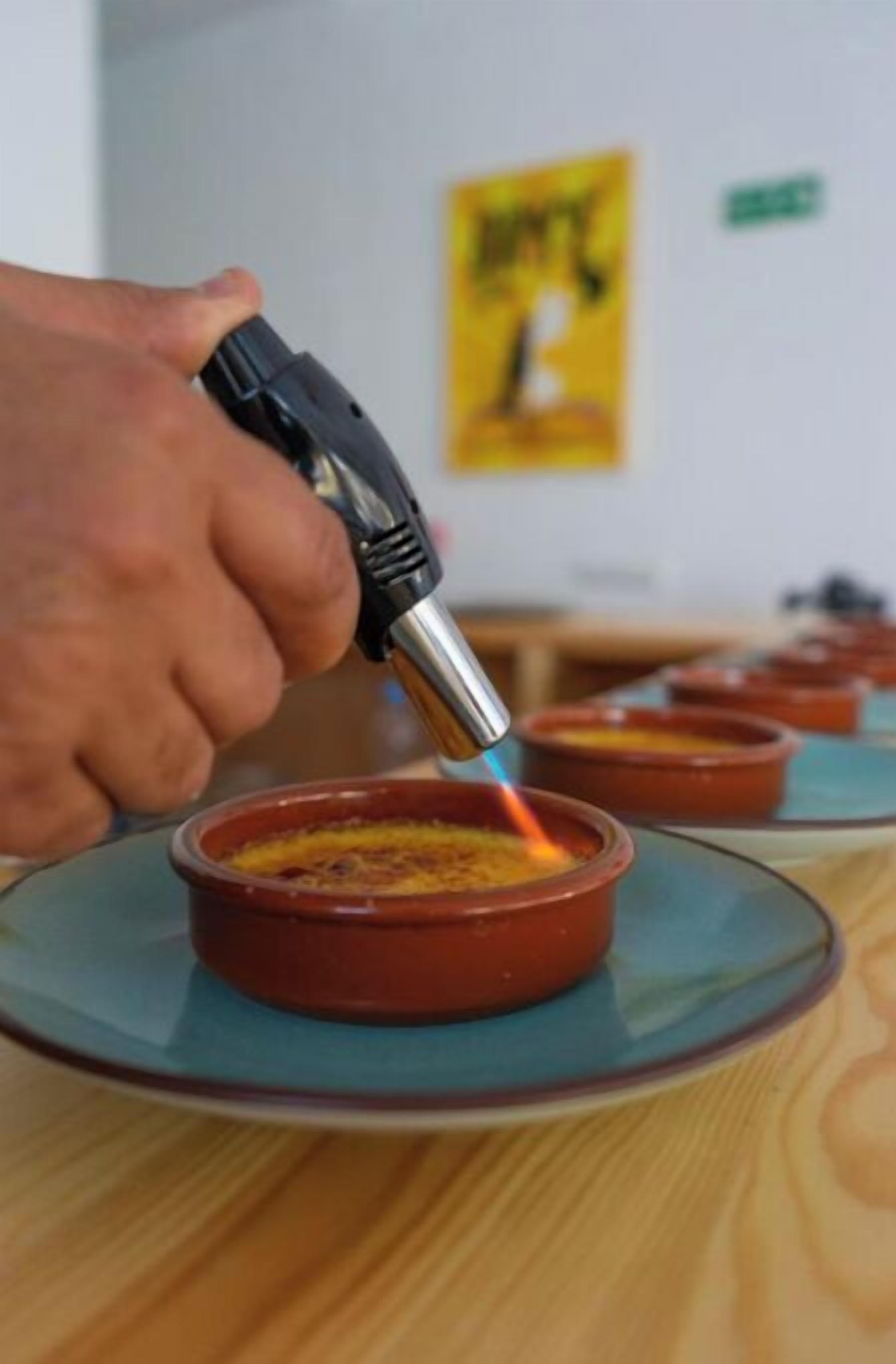 A hand holding a blowtorch over a dish, with several similar dishes lined up in the background.