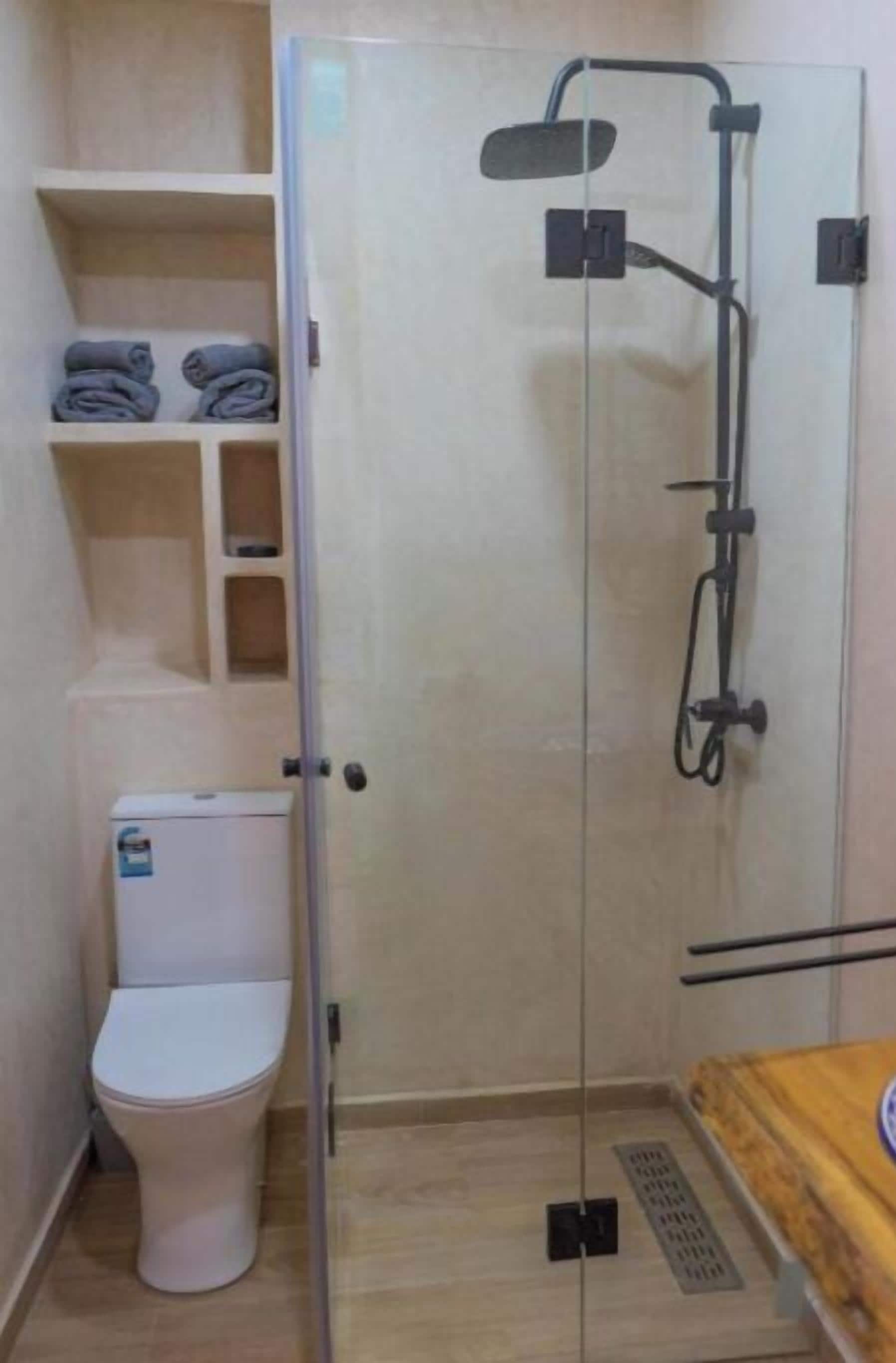 Un baño pequeño con inodoro, ducha con cerramiento de vidrio y repisas con toallas dobladas.