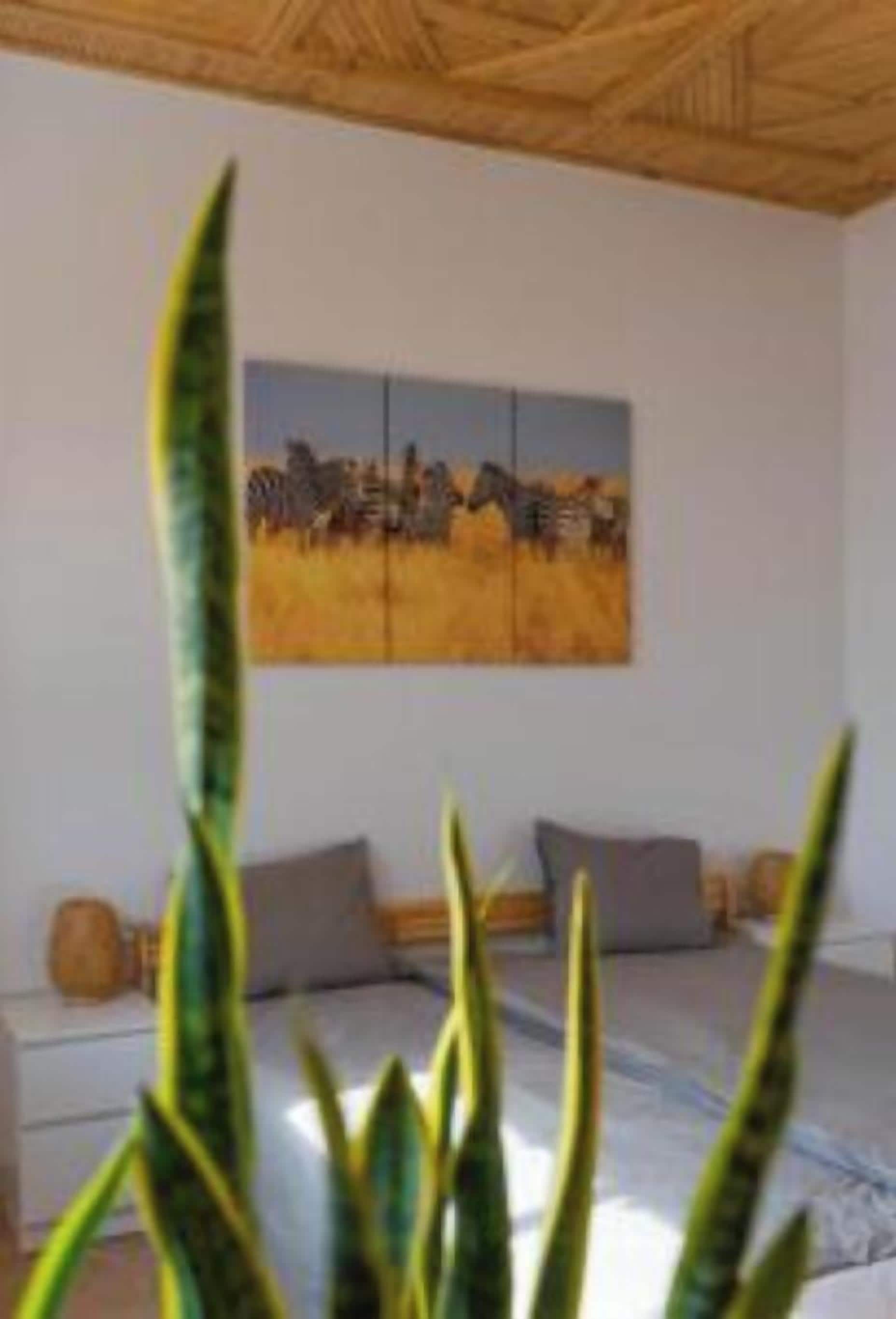 Una habitación con una cama, un cuadro de un campo con animales y una planta al frente.