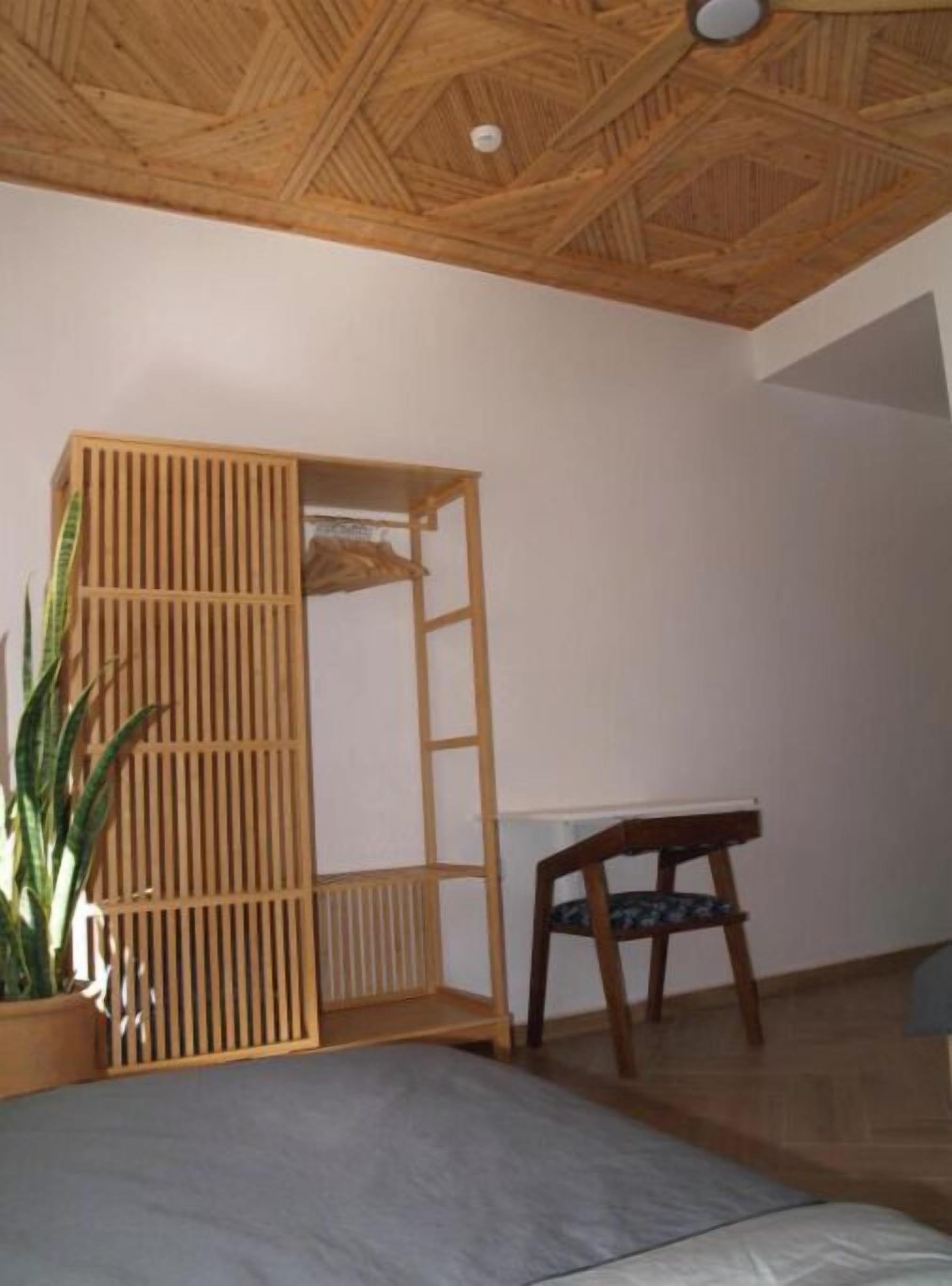 A room with a wooden ceiling, a bed, a desk, a chair, and a potted plant.
