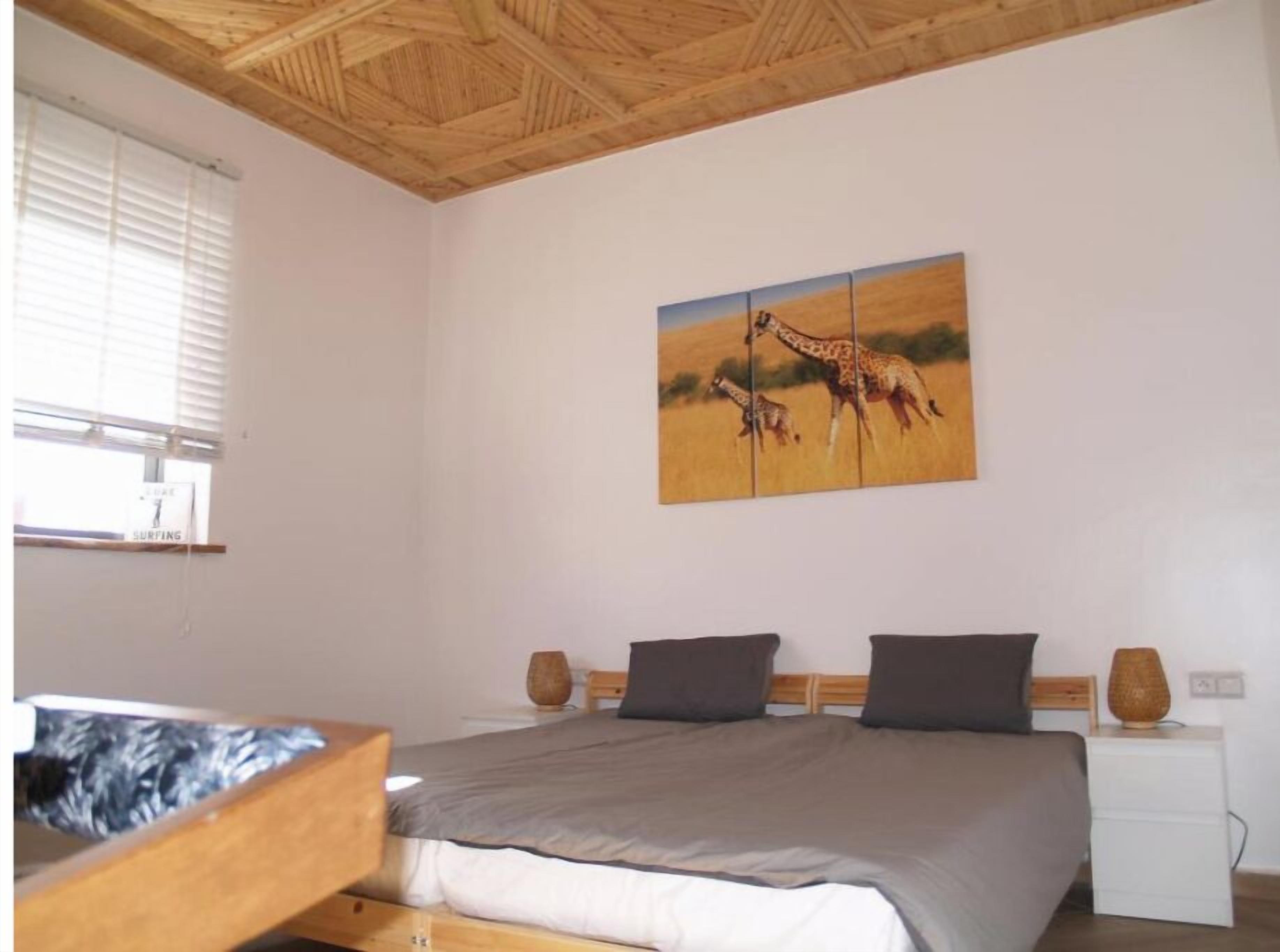 A bedroom with a bed, a painting of giraffes, a window with blinds, and a wooden ceiling.