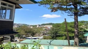 A residential building with a green roof, a large tree in front, and a view of a town in the background. - Baden Powell Hostel  (Baguio)