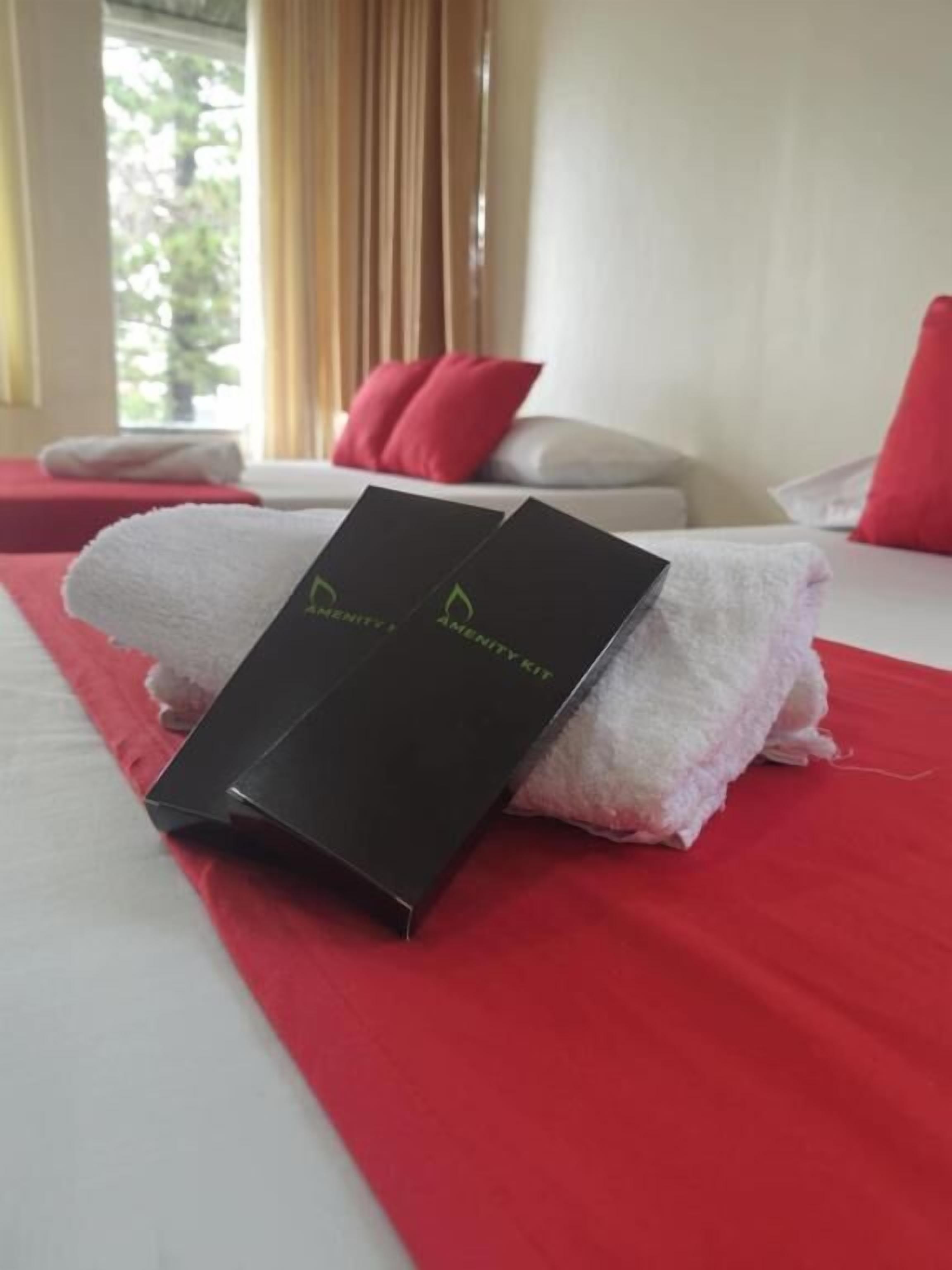 A neatly made bed with a red bedspread, a white towel, and a black box labeled "AMENITY KIT" on top.
