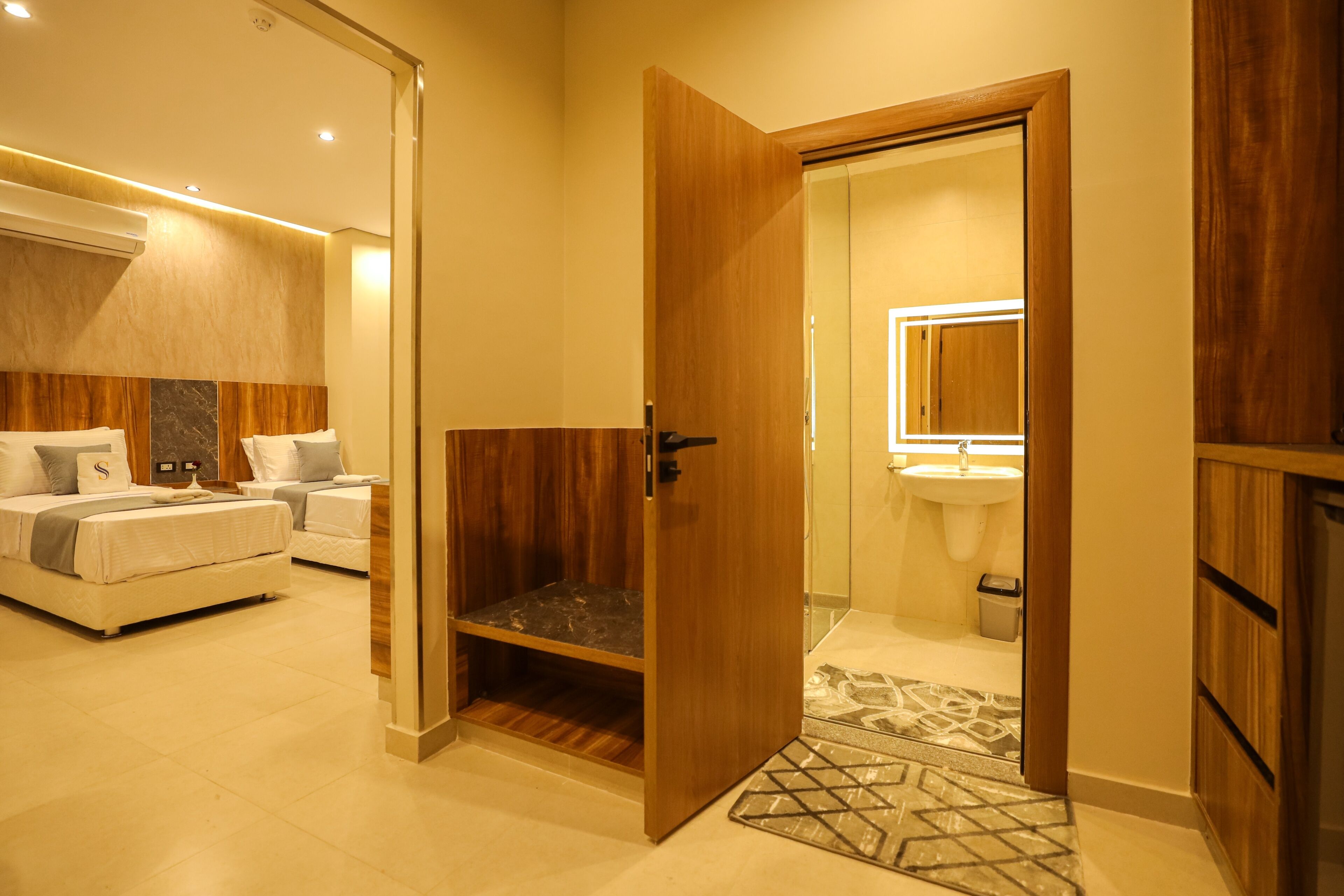 Une chambre d’hôtel moderne avec une armoire en bois, un lit avec une tête de lit, une table de chevet et une salle de bain équipée d’un lavabo et d’un miroir.