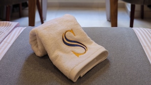 A neatly made bed with a towel featuring a monogram, a wooden table with a glass of orange juice, and a view of buildings through a window with curtains.