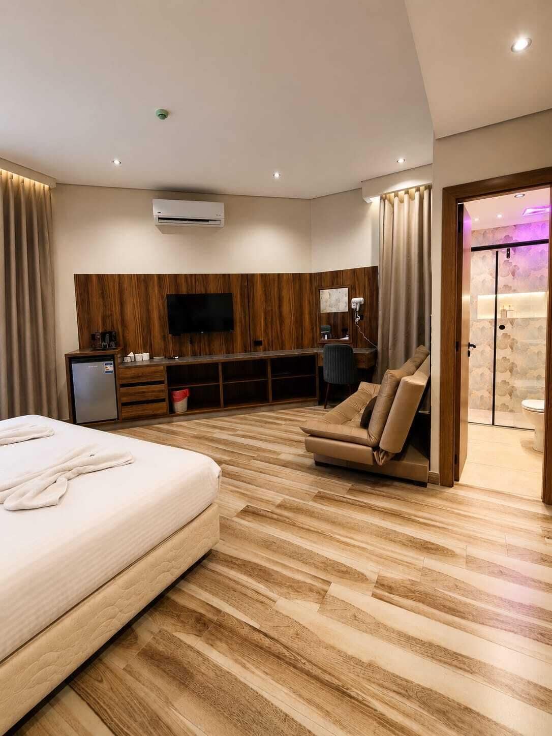 Habitación de hotel moderna con una cama grande, un sofá, un televisor montado en la pared y un baño visible a través de una puerta de cristal.