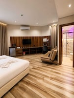 Habitación de hotel moderna con una cama grande, un sofá, un televisor montado en la pared y un baño visible a través de una puerta de cristal.